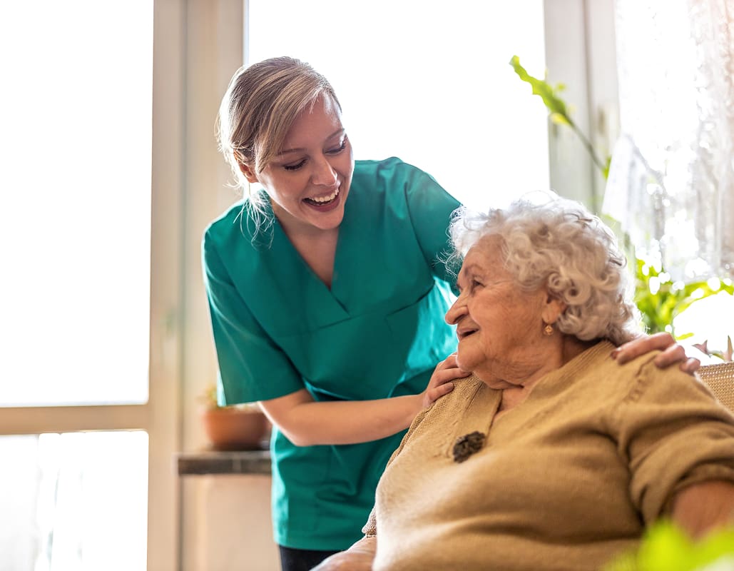A caregiver in teal scrubs gently touches an elderly woman's shoulder, both smiling warmly and making eye contact in a bright room with large windows and green plants.