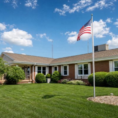 A single-story building with a brick exterior, surrounded by well-maintained green lawns and bushes. An American flag is flying on a tall flagpole in the foreground. The sky is blue with scattered white clouds.