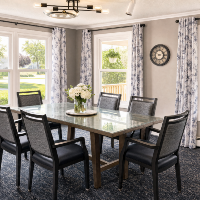 A dining room with a large rectangular table surrounded by eight black chairs. A vase of white flowers is on the table. There are three windows with floral curtains, allowing natural light into the room. A round wall clock is on the wall to the right. The ceiling has a modern light fixture.