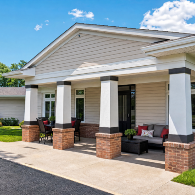 A covered porch with four white columns and brick bases, featuring outdoor seating including a black sofa with red and white pillows, and a black table with chairs, in front of a light-colored building under a blue sky with some clouds.
