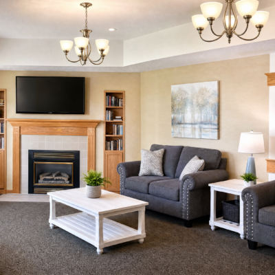A cozy living room with a fireplace, a flat-screen TV above it, and a wooden bookshelf to the right. There is a dark gray sofa with two pillows, a white coffee table with a small green plant, and a side table with a lamp and another green plant. Two chandeliers hang from the ceiling.