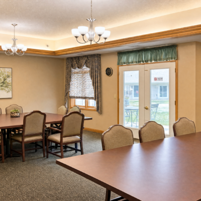 A dining area with multiple tables and chairs, featuring a window with patterned curtains, a door leading outside, and ceiling lights providing illumination.