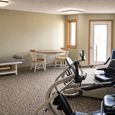 A room with beige walls and carpet, furnished with exercise equipment, a small round table with four wooden chairs, a window, a door with a window, and a padded bench with a pillow.