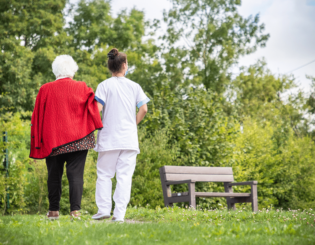 An elderly woman with white hair and a woman in a white uniform walk together on a grassy area with trees in the background. There is a wooden bench nearby.