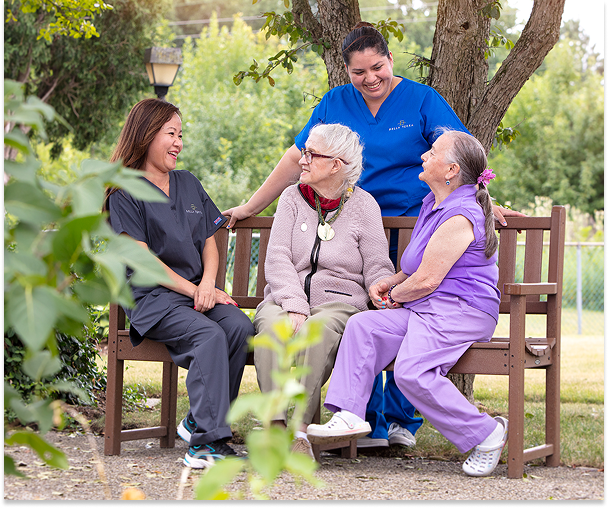 Four women, including two elderly women and two caregivers, are sitting and standing around a wooden bench outdoors, smiling and engaging in conversation. One elderly woman wears glasses and a pink sweater, the other wears purple scrubs, and the caregivers wear navy blue uniforms. They are surrounded by greenery and trees.