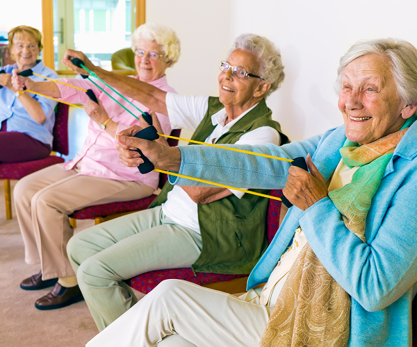 Four elderly women are seated in a row, participating in a seated exercise class using resistance bands. They are smiling and appear engaged in the activity.