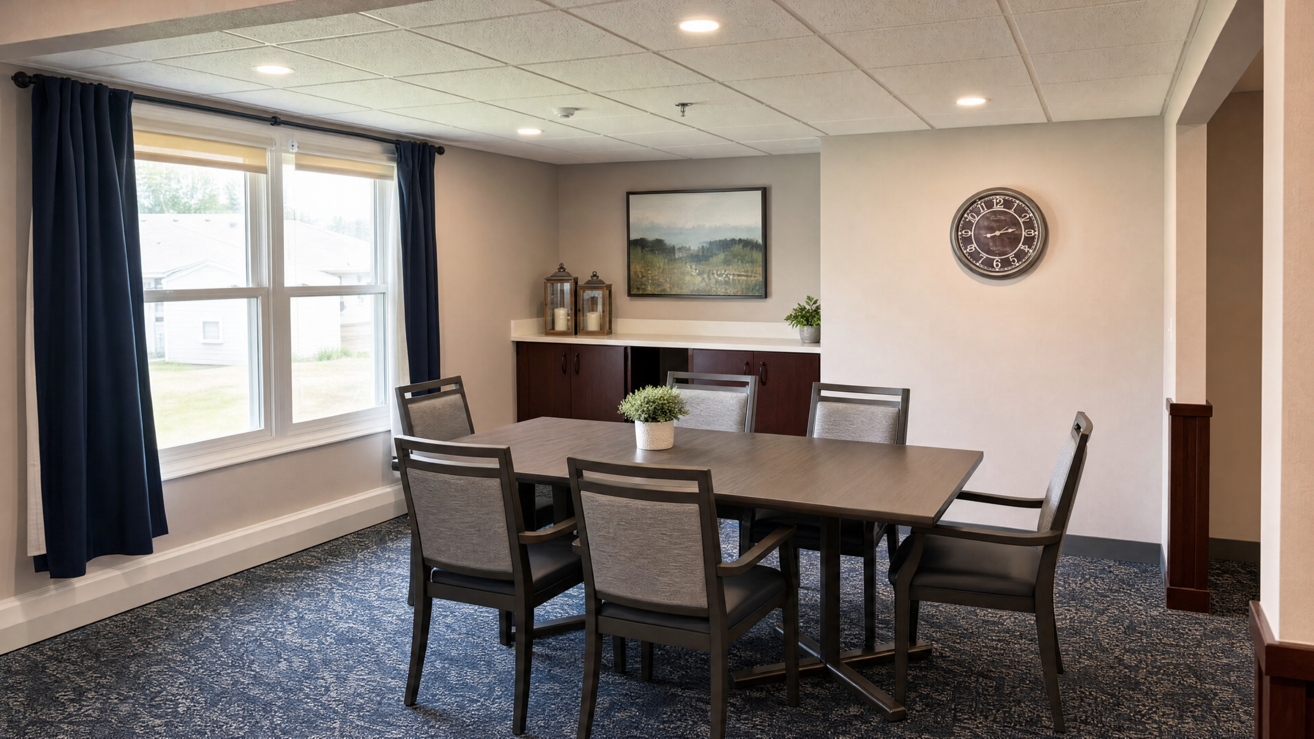 A dining area with a rectangular table surrounded by six gray upholstered chairs. There is a potted plant in the center of the table, a large window with blue curtains on the left, and a sideboard with decorative lanterns, a painting, and a potted plant on the back wall. A round wall clock is mounted on the right wall. The room has a neutral color scheme with beige walls and a textured carpet.