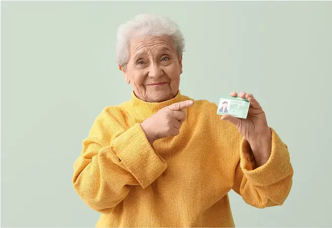 An elderly woman with white hair, wearing a yellow sweater, smiling and pointing to her driver's license she holds in her right hand against a light green background.