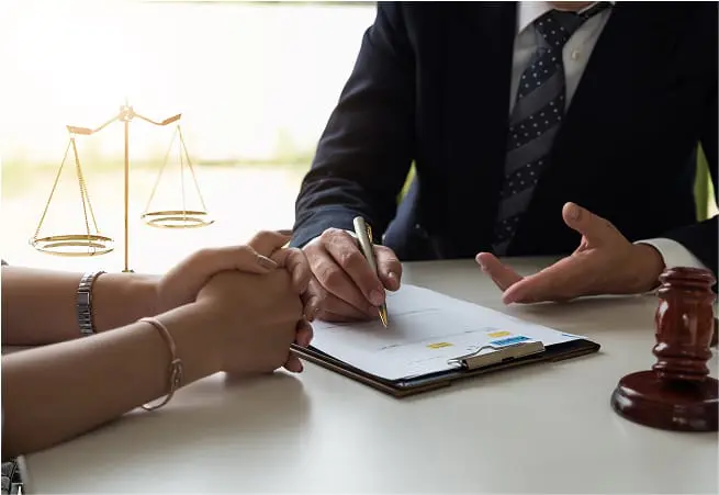 A person in a suit is signing a document at a table, while another person's hands are folded and resting on the table. A set of scales and a gavel are also visible on the table.
