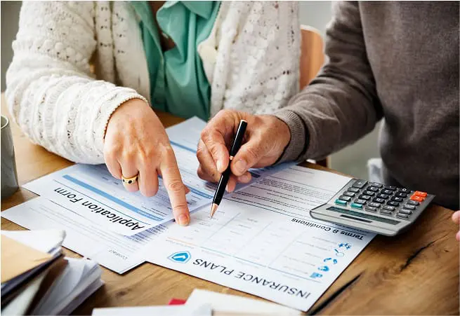Two people are sitting at a table reviewing documents, including an insurance plan and what appears to be a form; one person is pointing at the documents while the other is holding a pen and a calculator.