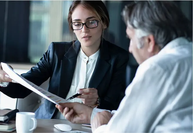 A young woman in business attire and glasses discusses documents with an older man in a white shirt, sitting at a table in a professional setting.