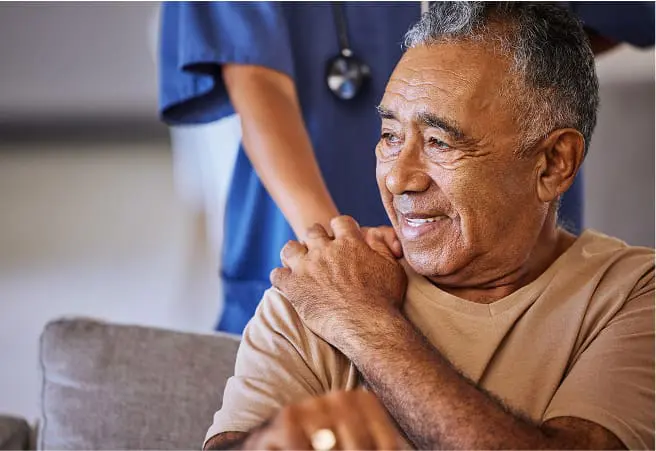 An elderly man sitting on a couch, smiling, with a healthcare worker standing behind him. The healthcare worker is wearing blue scrubs and a stethoscope around their neck. The man has his left arm crossed over his chest, with his hand resting on his shoulder.