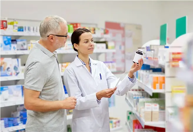 A female pharmacist shows a bottle of medication to an older male customer inside a pharmacy, with shelves of medicines and health products in the background.