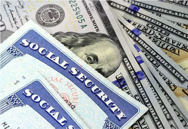 A close-up of U.S. currency, including Social Security cards and various dollar bills, with a focus on the Social Security cards in the foreground and dollar bills partially visible behind.