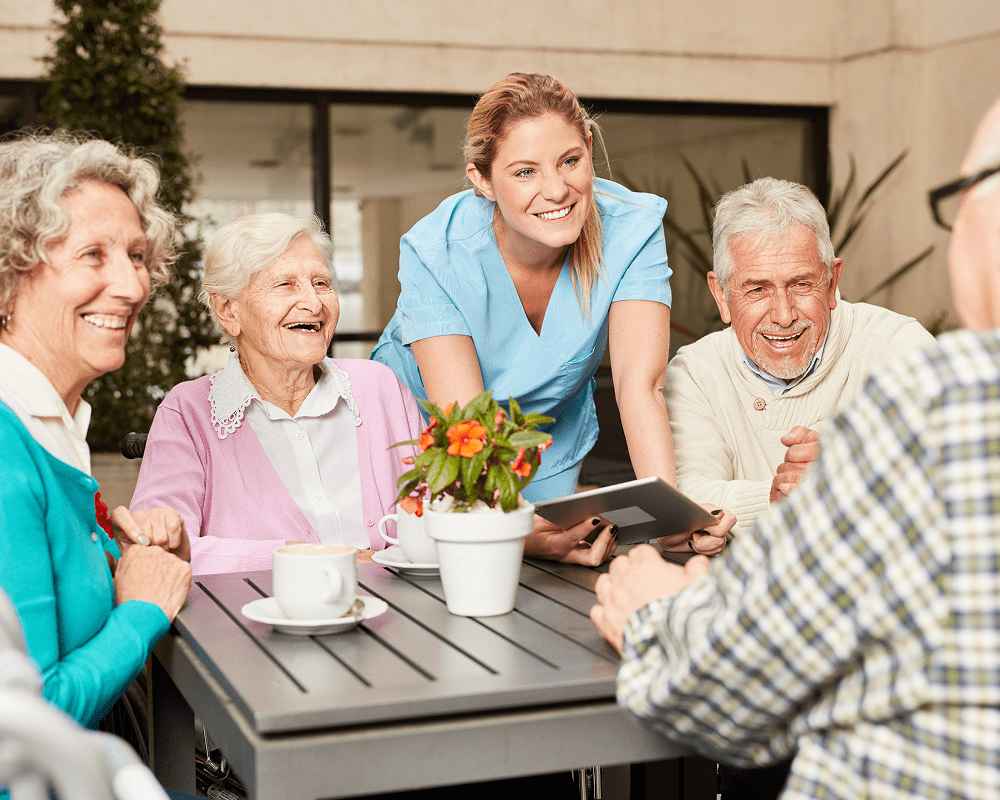 A group of elderly people sitting around a table outdoors, smiling and engaging with a young female caregiver who is showing them a tablet. There are cups of coffee and a potted plant on the table.